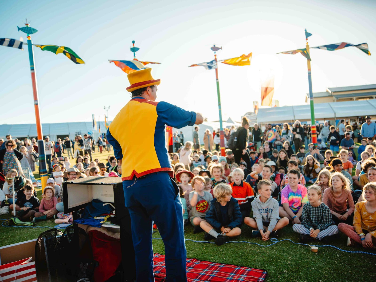 A circus performer with a yellow and blue top on, and a yellow hat performs a crowd of smiling kids.
