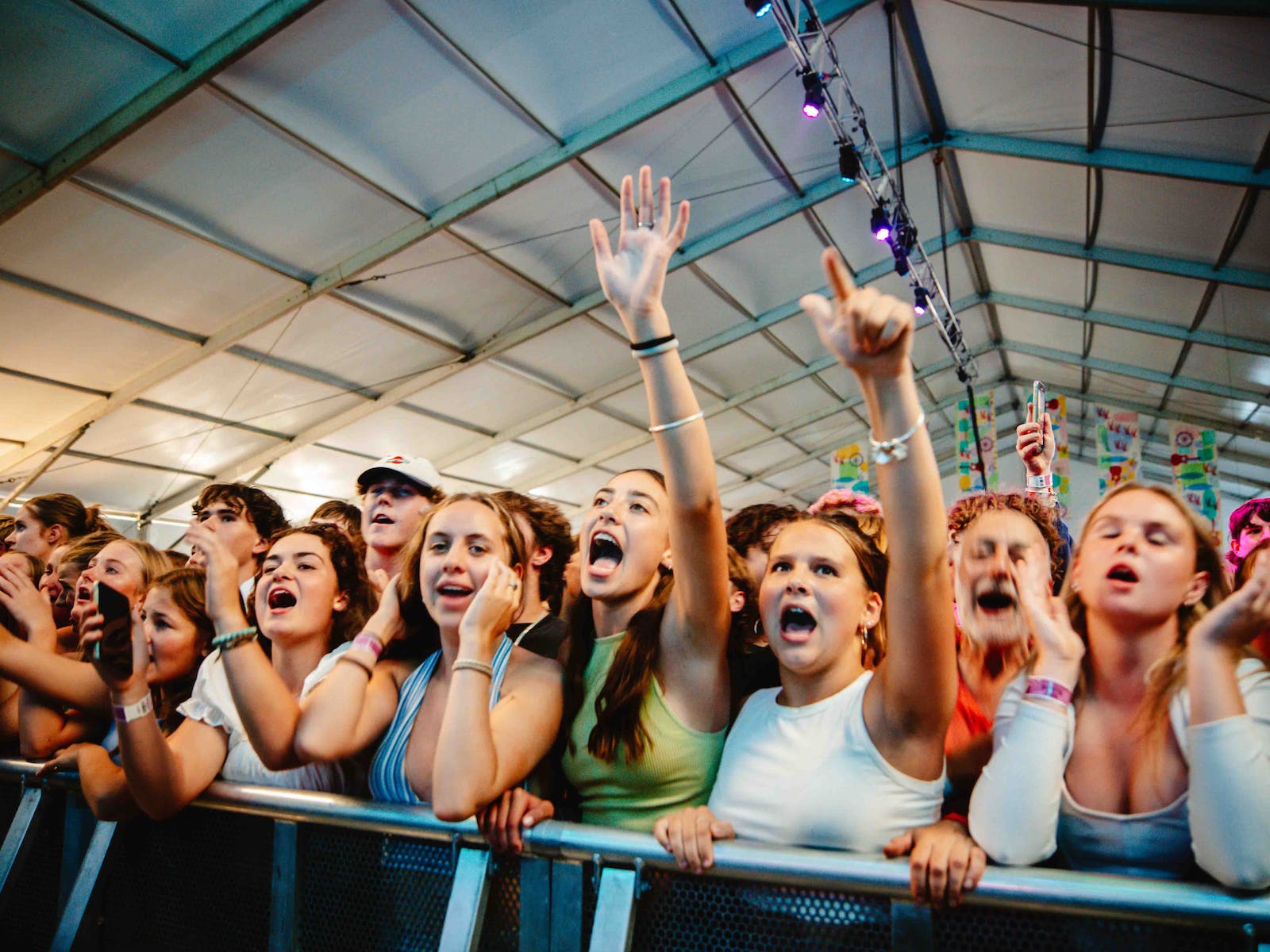 Young people sing along to music against a fence inside a tent. Two people have their arms up.