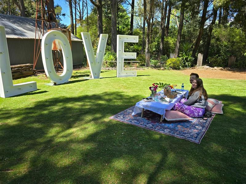 Photo of a man and woman sitting on a rug layed on a grassed area. A large love sign and picnic