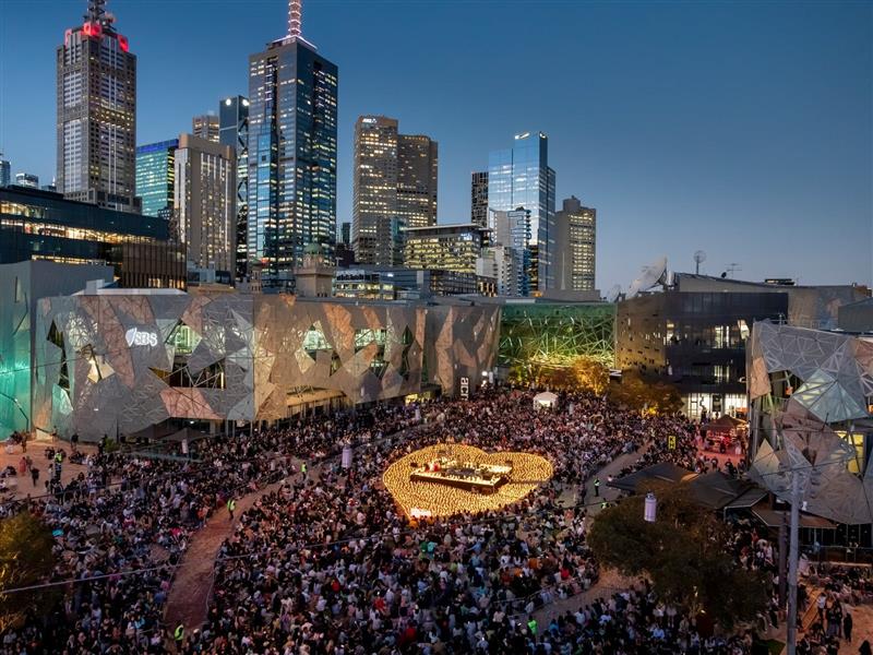 Thousands gather in Fed Square around a stage, surrounded by candles in the shape of a heart.