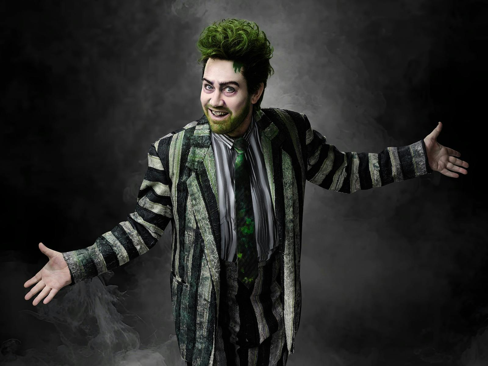 A man stands in a misty spotlight with his arms outstretched. He is wearing a striped suit.